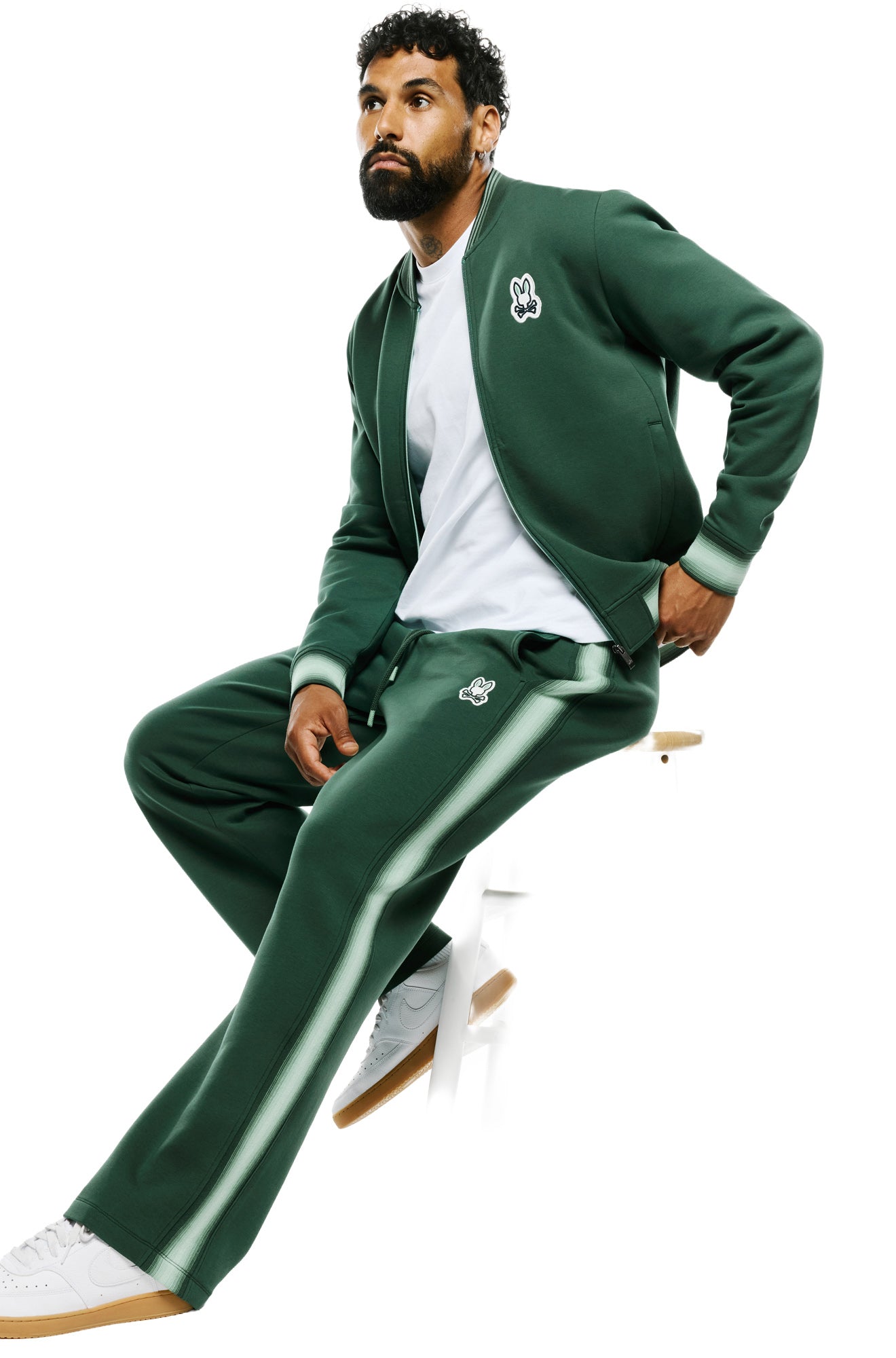 Psycho Bunny men's angled seated view of dark green sweatpants showing thigh logo patch and the light green side stripe