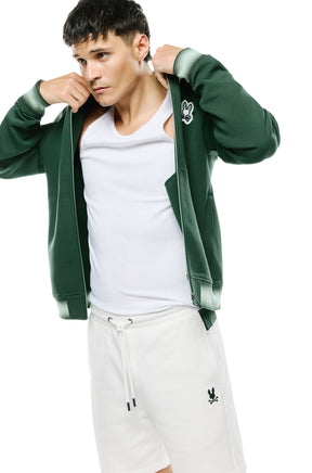 Men's angled model view of open dark green bomber jacket showing striped cuffs, pocket and zipper, side view by Psycho Bunny