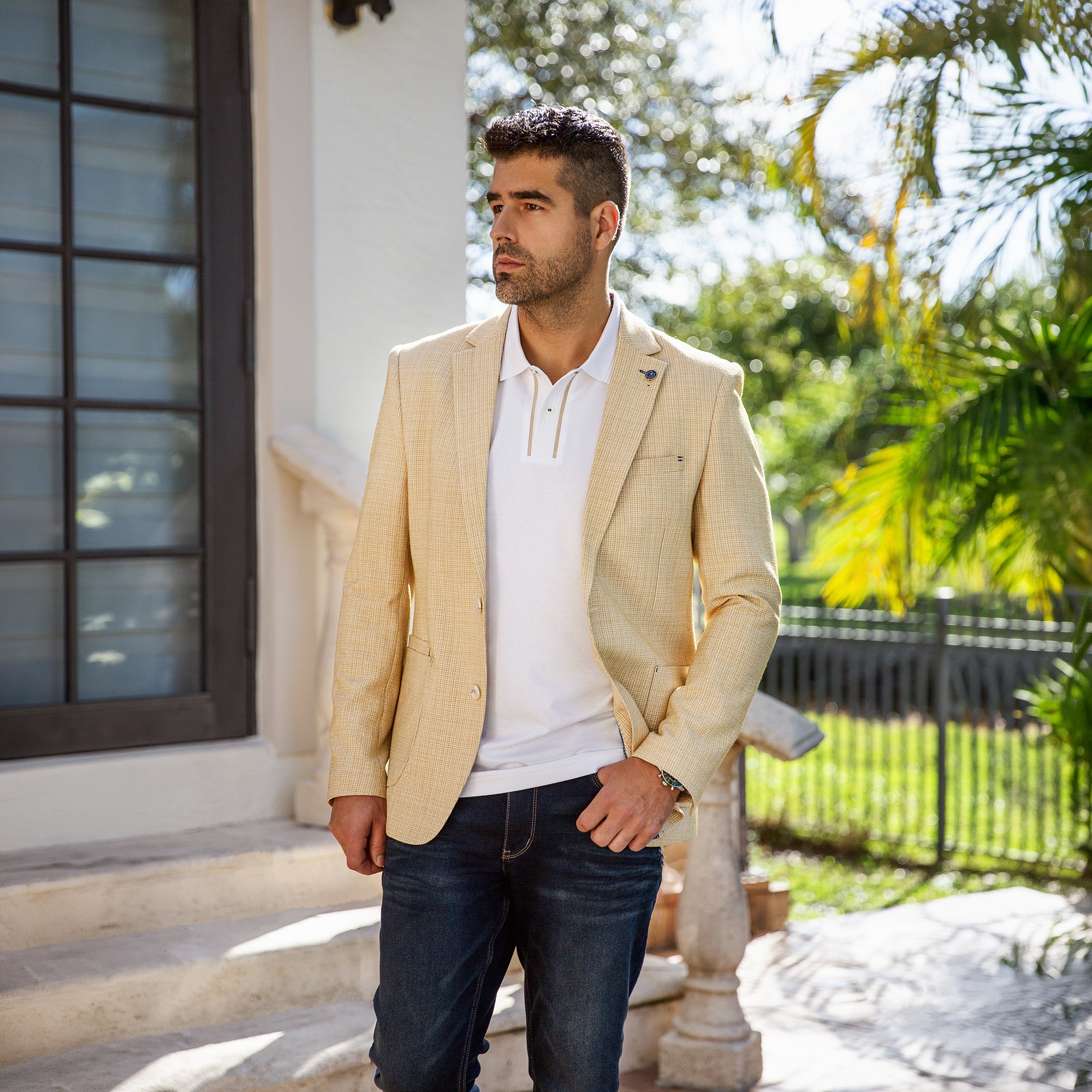 Veston beige Au Noir pour homme porté avec polo blanc et jeans foncés, dans un décor extérieur ensoleillé et chic.