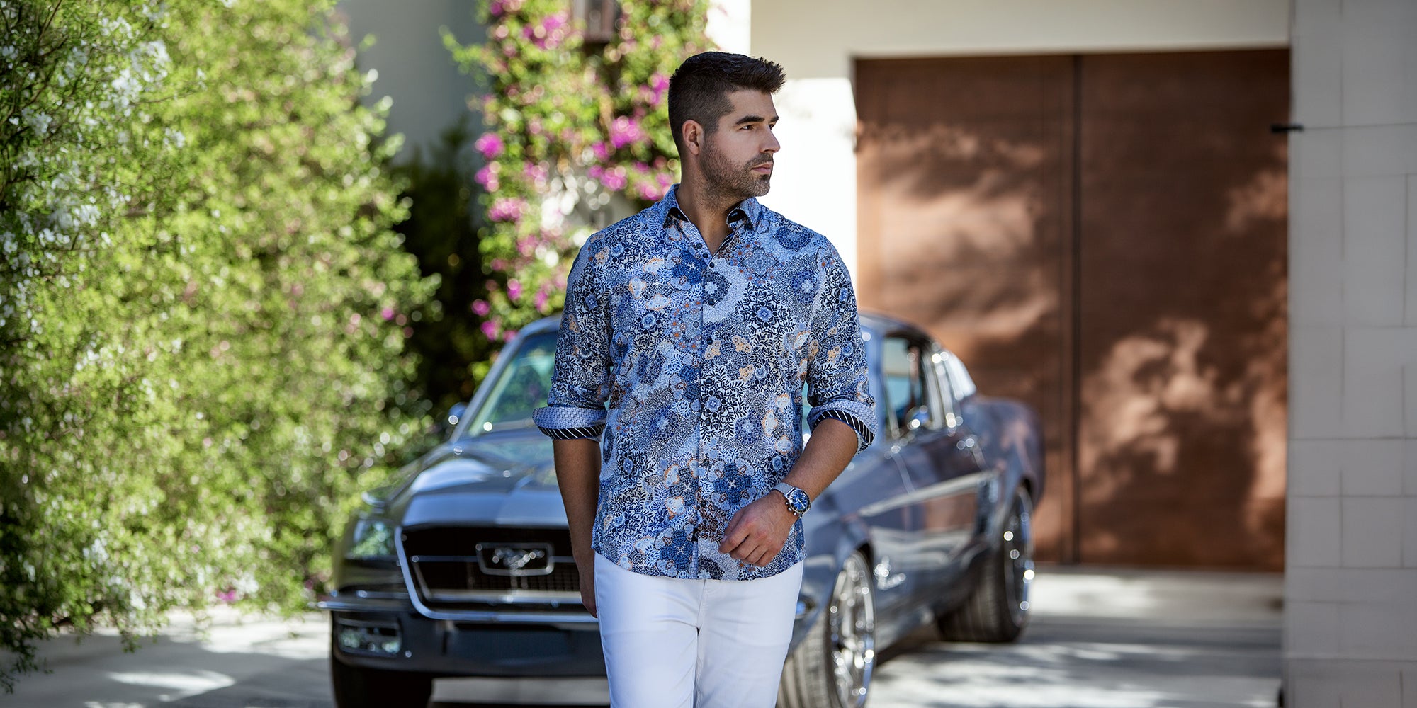 Chemise habillée bleue Au Noir pour homme à motif floral, portée avec pantalon blanc dans un décor extérieur élégant avec voiture classique.